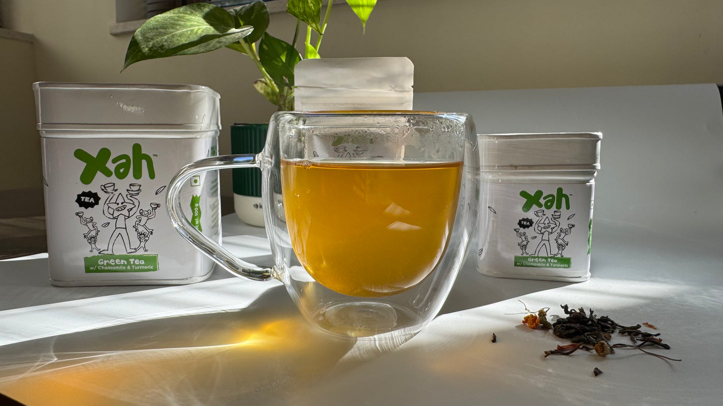 Clear glass mug with tea, Xah branded containers, and dried tea leaves on a light surface.