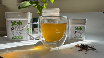 Clear glass mug with tea, Xah branded containers, and dried tea leaves on a light surface.